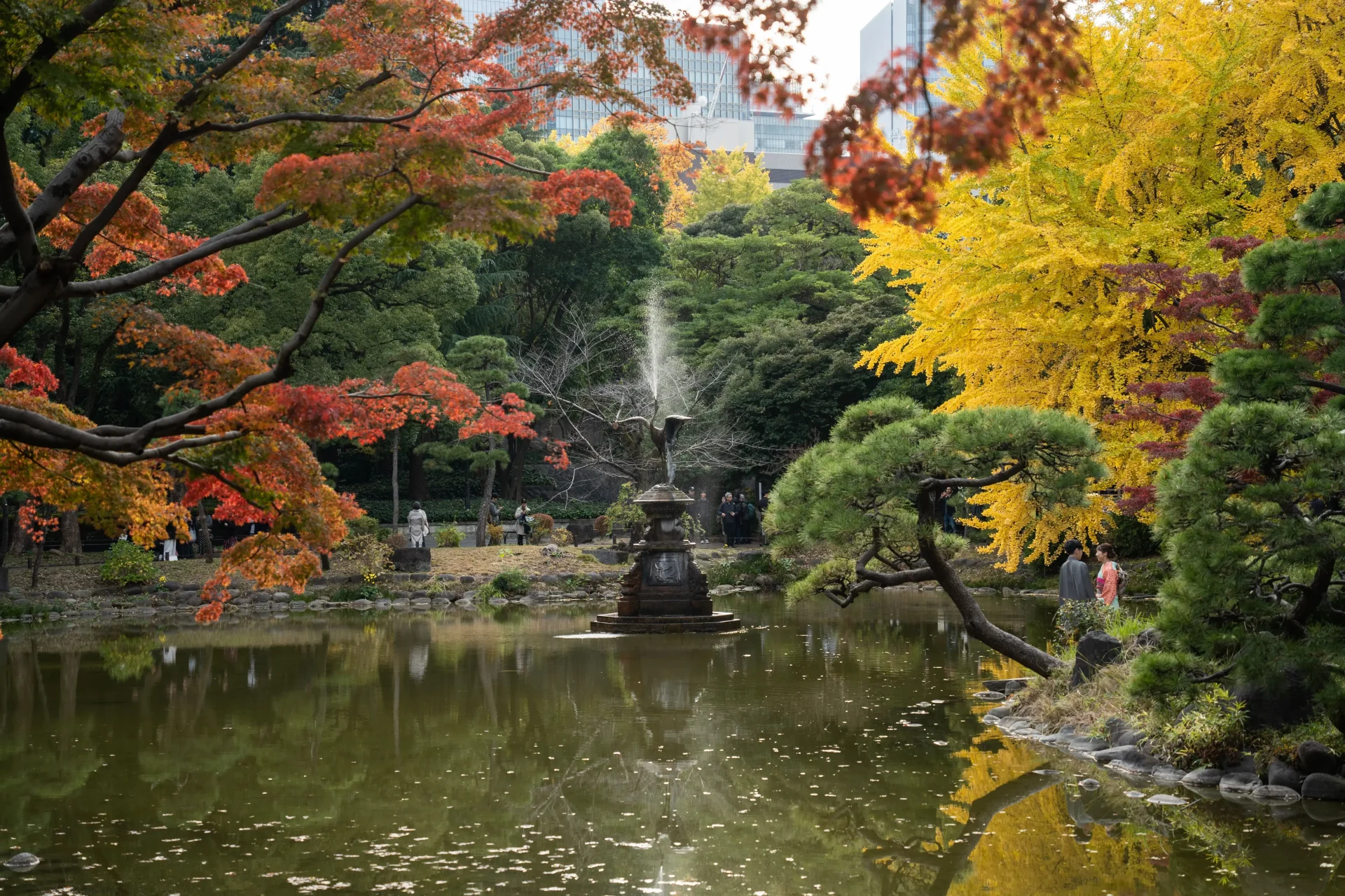 A traditional Japanese garden pond in a Tokyo park, reflecting vibrant crimson and orange autumn foliage with a small wooden bridge in the background.
